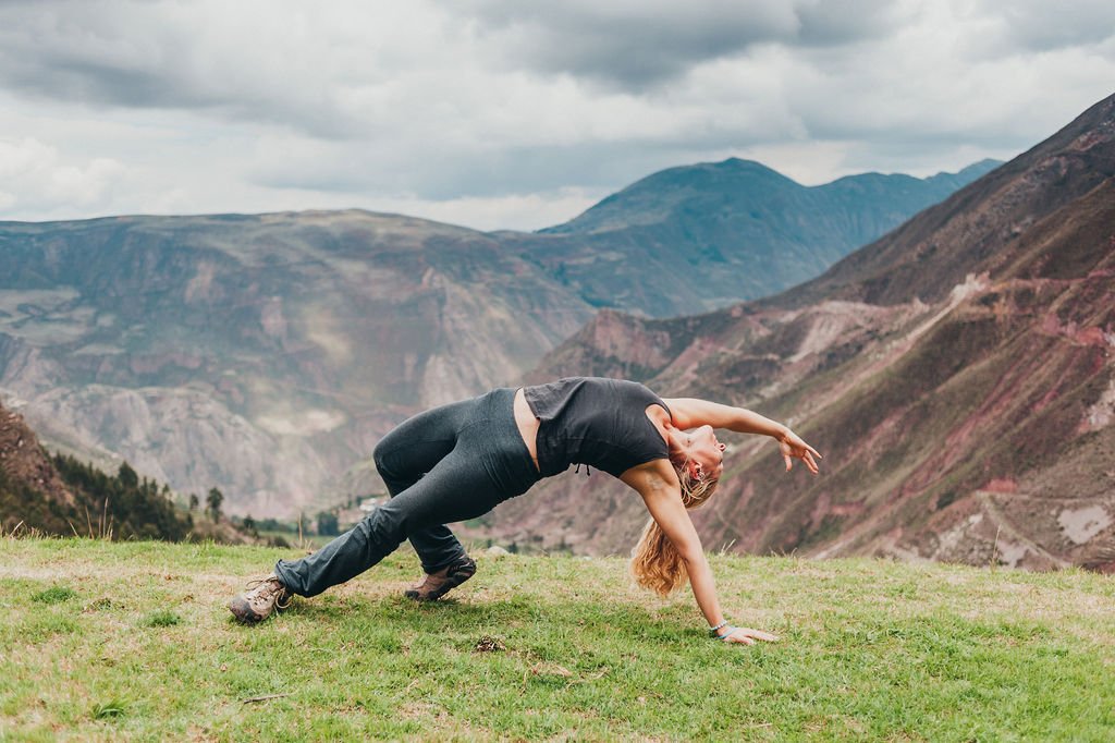 A woman performs a yoga backbend on a grassy hilltop overlooking the dramatic mountainous landscape of the Sacred Valley.