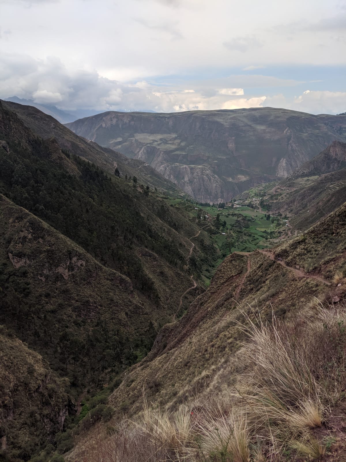 A high-angle view looks down into a deep green valley with a winding dirt path on a steep hillside in the Andes of Peru's Sacred Valley.
