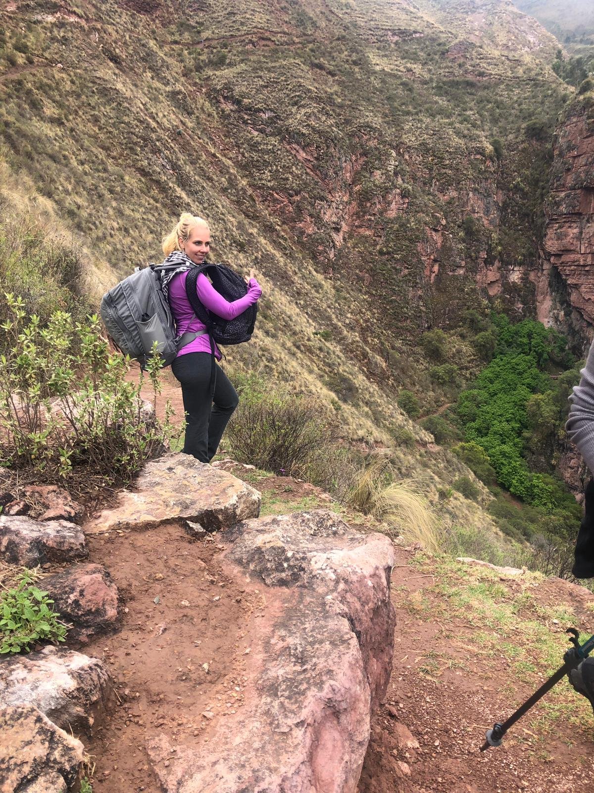 A woman hiker in a purple shirt gives a thumbs up on a steep mountain trail overlooking a lush canyon in the Sacred Valley.
