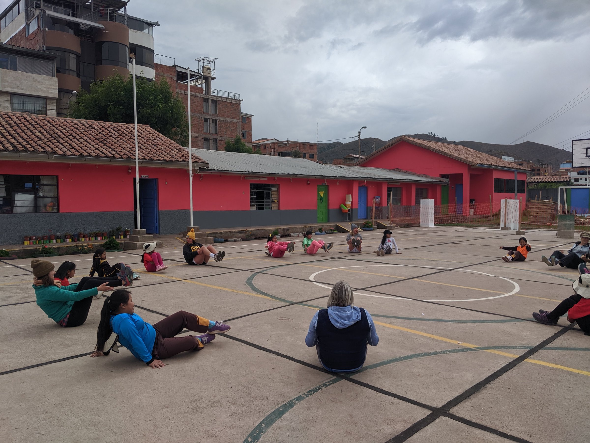 Retreat participants and local children sit in a large circle on an outdoor court practicing a core-strengthening exercise in front of a pink building in Cusco.