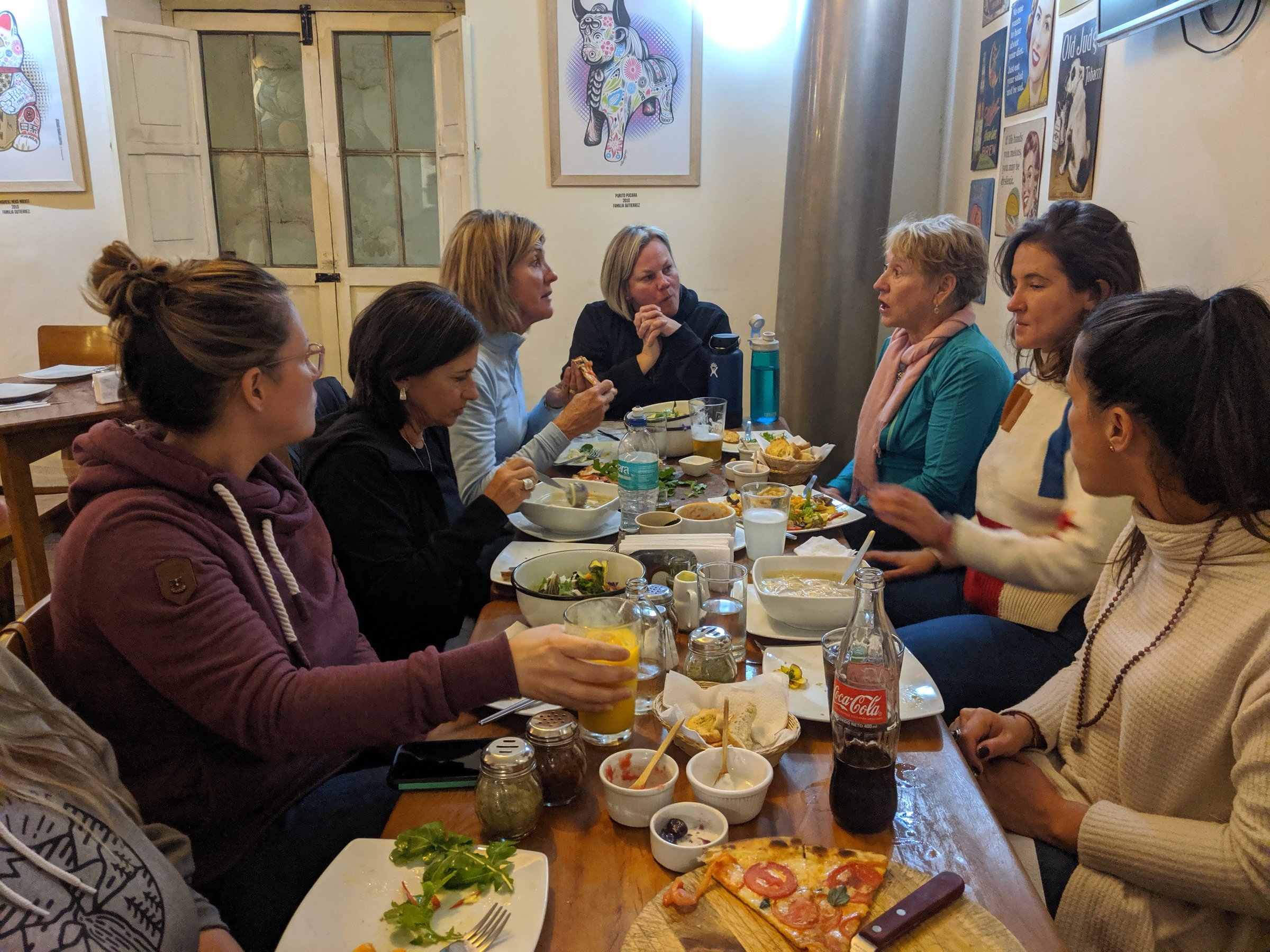 A group of women share a meal and conversation around a crowded wooden table at a restaurant in Cusco.