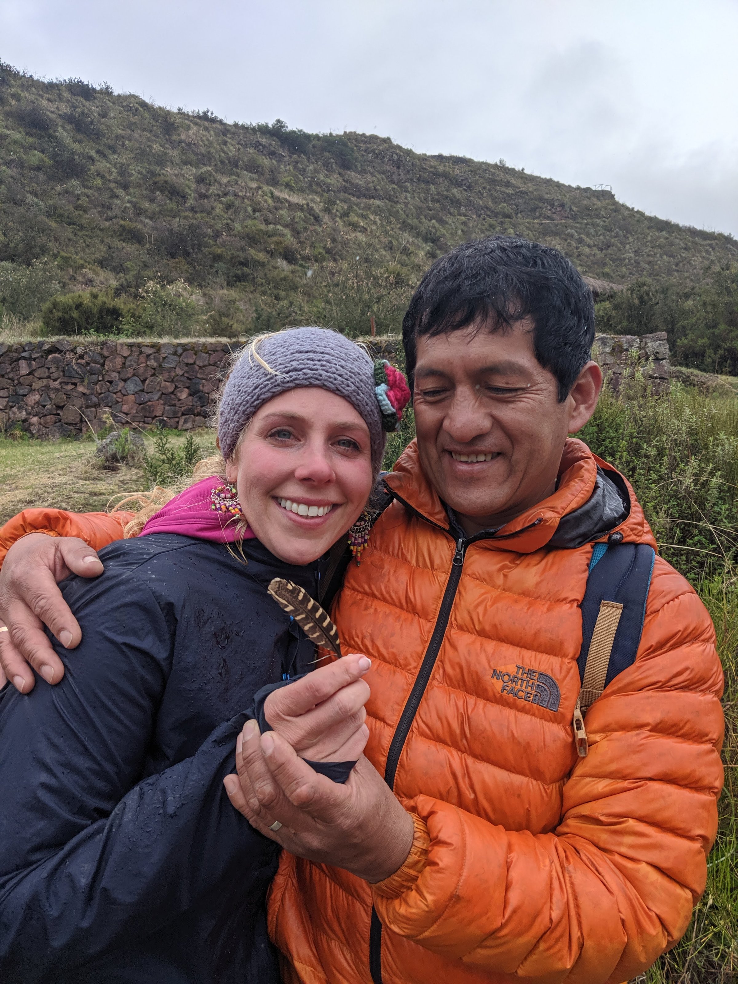 A woman in a grey headband and a man in an orange puffer jacket smile while holding a small patterned feather together on a grassy hillside in the Andes.