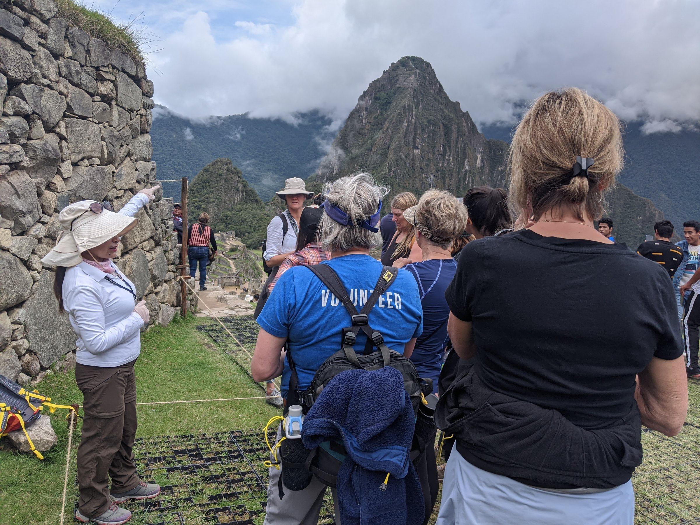 A tour guide points toward the misty peak of Huayna Picchu while a group of visitors listens at the Machu Picchu archaeological site.